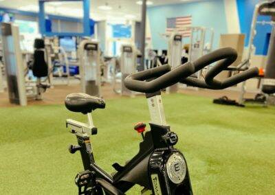 Exercise bike in foreground with gym equipment inside a fitness center with blue and white walls.