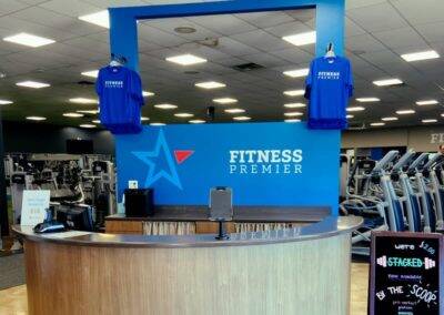 Front desk of fitness premier gym with blue branding, staffed computers, and hand sanitizers, surrounded by exercise equipment.