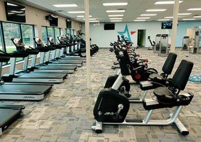 A modern gym with rows of treadmills and stationary bikes overlooking windows, accompanied by other fitness equipment in a clean, organized space.