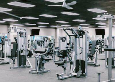 Modern gym interior with rows of various exercise machines and ceiling fans, captured in a cool-toned monochrome color scheme.